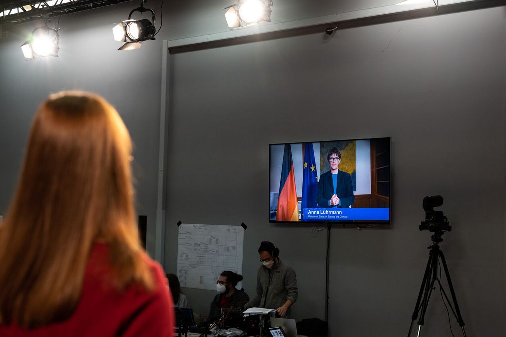 zur Vergrößerungsansicht des Bildes: Frau mit Rücken zur Kamera schaut auf Bildschirm, auf dem eine Frau neben Europa und Deutschlandflagge spricht