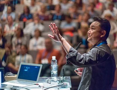 Ein Mann asiatischer Herkunft steht in einem Hörsaal und hält einen Vortrag. Im Hintergrund sind die Zuhörer abgebildet. Im Vordergrund steht der Vortragende mit einem Laptop und Mikrofon.