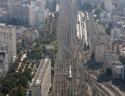 Luftbild des Pariser Bahnhofs Gare Montparnasse. Ein Geflecht von zahlreichen Bahngleisen zieht sich über die gesamte Bildhöhe. Links und rechts der Gleise stehen dicht aneinander Wohngebäude und Hochhäuser, die zusammen ein beeindruckendes Bild einer dicht bevölkerten Metropole hinterlassen.