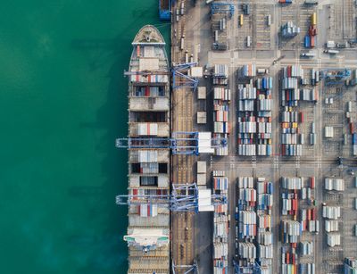 Luftansicht auf einen Frachthafen. Rechts im Bild lagern hunderte Container, links im Bild wird ein großes Containerschiff beladen.