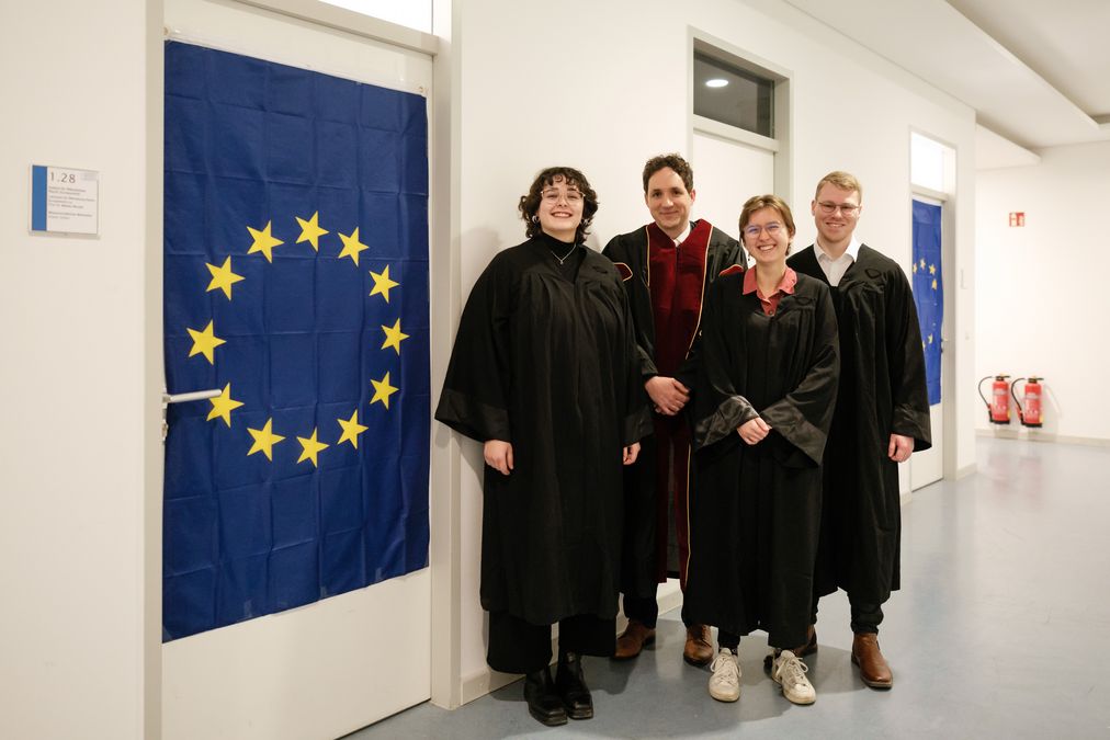 zur Vergrößerungsansicht des Bildes: Vier Personen stehen neben Europaflagge und lächeln in Kamera