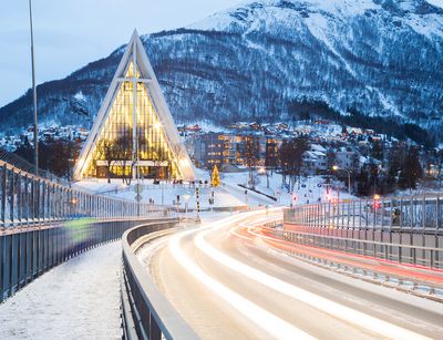 Blick in der Dämmerung auf die hell erleuchtete Arctic Cathedral im norwegischen Tromsø. Im Hintergrund sind ein schneebedeckter Berg sowie am Fuße des Berges erleuchtete Wohnhäuser sowie die Kirche zu sehen. Im Vordergrund führen eine Straße und daneben ein Fußweg über eine Brücke am Berg vorbei. Auf dem Bild wurden die Lichtspuren der Fahrzeuge festgehalten.